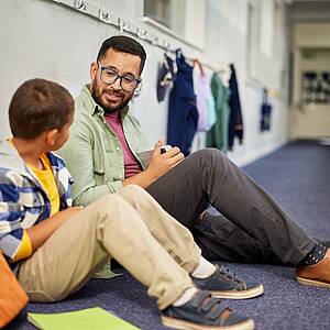 Lehrer spricht mit einem Jungen, sie sitzen auf dem Boden im Gang vor den Klassen
